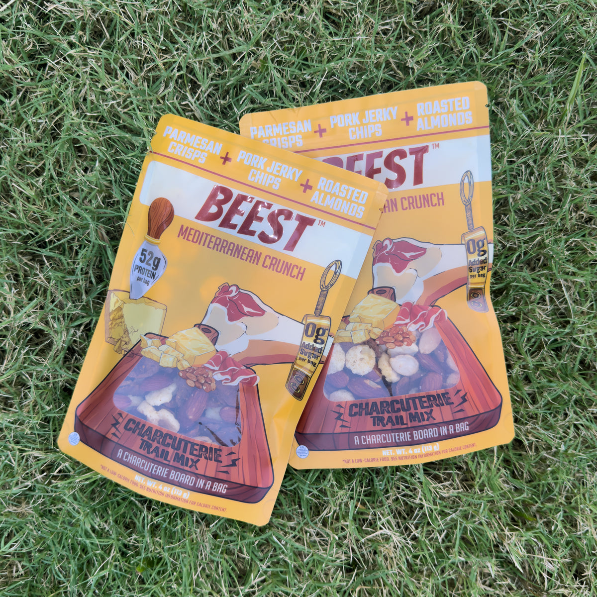 Two bags of BEEST Charcuterie Trail Mix, Mediterranean Crunch, crossed on green grass