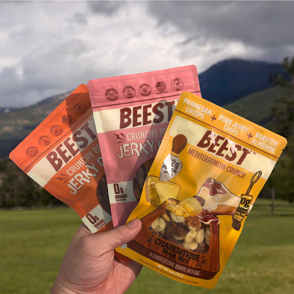 A hand holding The Herd: Charcuterie Trail Mix, Beef Jerky Chips and Pork Jerky Chips in a mountain meadow