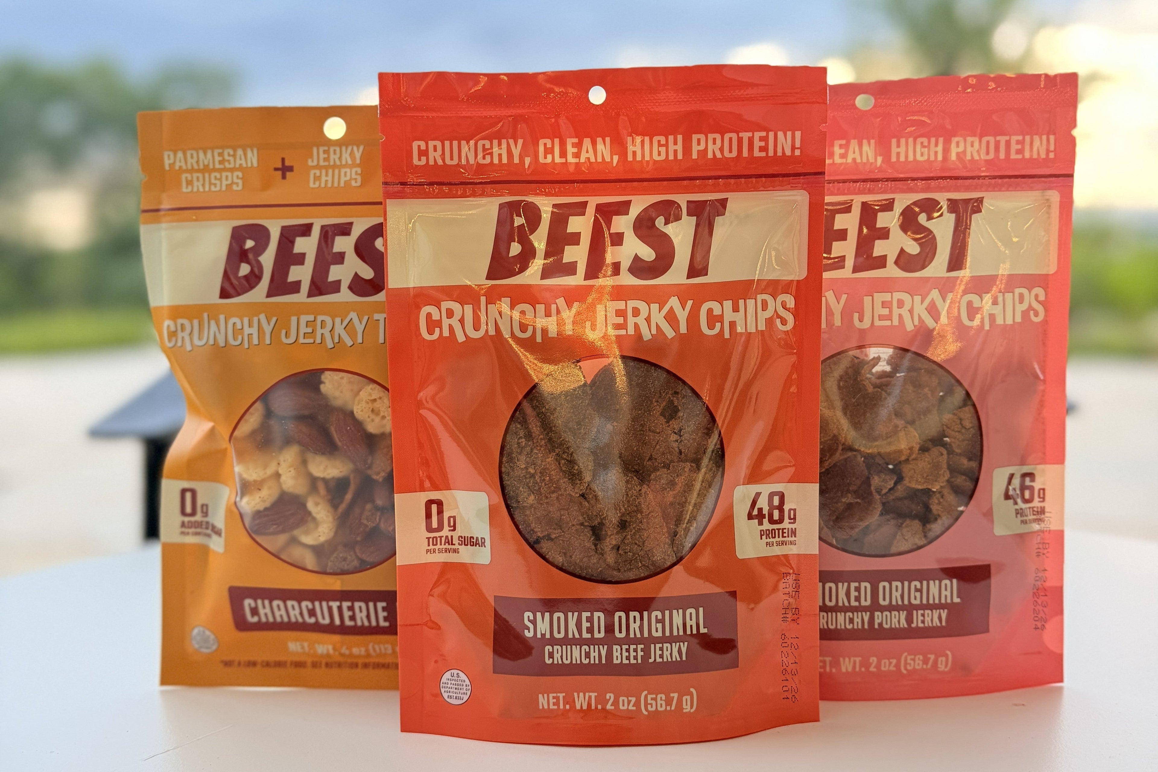 Three packages of BEEST Crunchy Jerky Chips on a blurred outdoor background