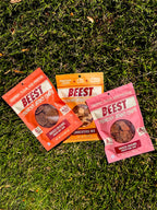 Three packages of BEEST Snacks. Crunchy Beef Jerky Chips, Charcuterie Trail Mix, and Crunchy Pork Jerky Chips, laying on grass. With claims shining in the sun, 48g of protein 0g Sugar, 52g of protein, 46 g of protein.