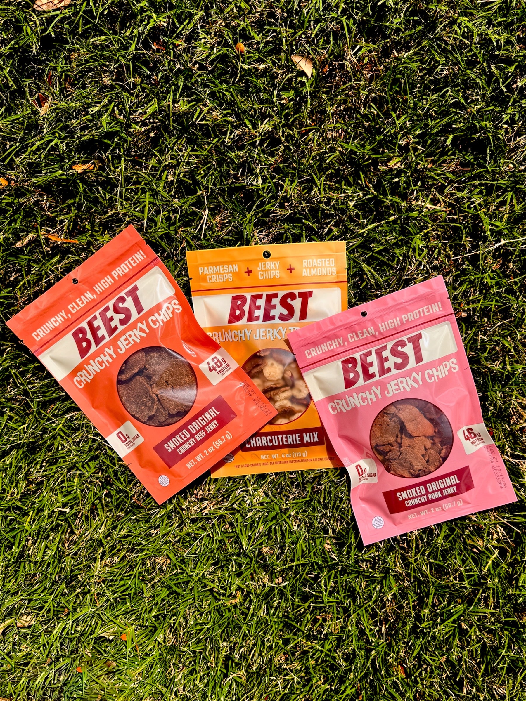 Three packages of BEEST Snacks. Crunchy Beef Jerky Chips, Charcuterie Trail Mix, and Crunchy Pork Jerky Chips, laying on grass. With claims shining in the sun, 48g of protein 0g Sugar, 52g of protein, 46 g of protein.