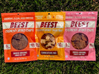 Three packages of BEEST Snacks. Crunchy Beef Jerky Chips, Charcuterie Trail Mix, and Crunchy Pork Jerky Chips, laying on grass. With claims shining in the sun, 48g of protein 0g Sugar, 52g of protein, 46 g of protein.