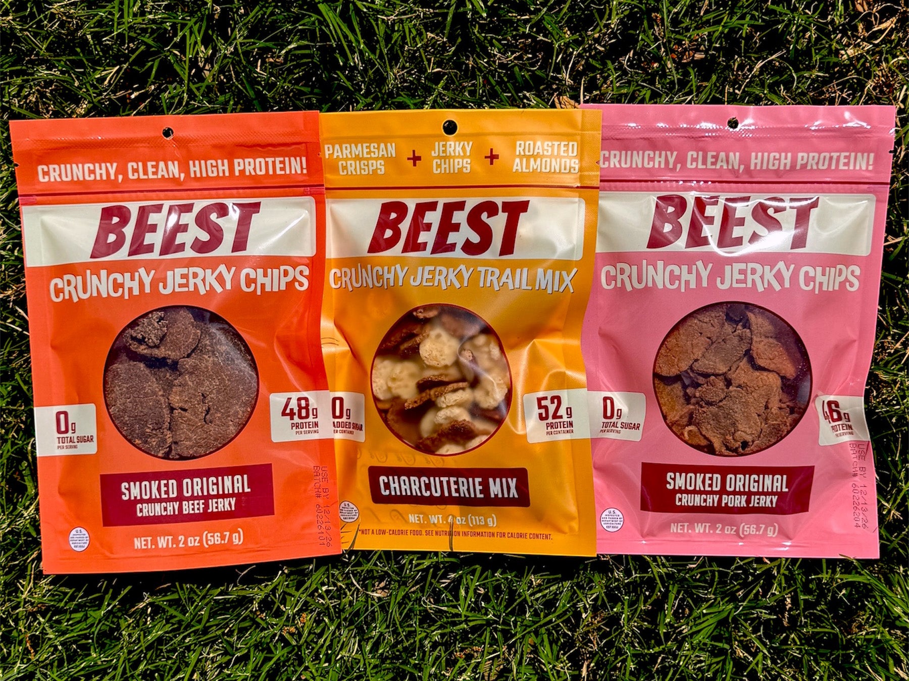 Three packages of BEEST Snacks. Crunchy Beef Jerky Chips, Charcuterie Trail Mix, and Crunchy Pork Jerky Chips, laying on grass. With claims shining in the sun, 48g of protein 0g Sugar, 52g of protein, 46 g of protein.