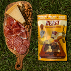 A bag of BEEST Mediterranean Crunch charcuterie trail mix, called "a charcuterie board in a bag" with 52g of protein and 0g added sugar, placed on green grass next to a wooden CHARCUTERIE board with cheese, salami, prosciutto, and almonds.